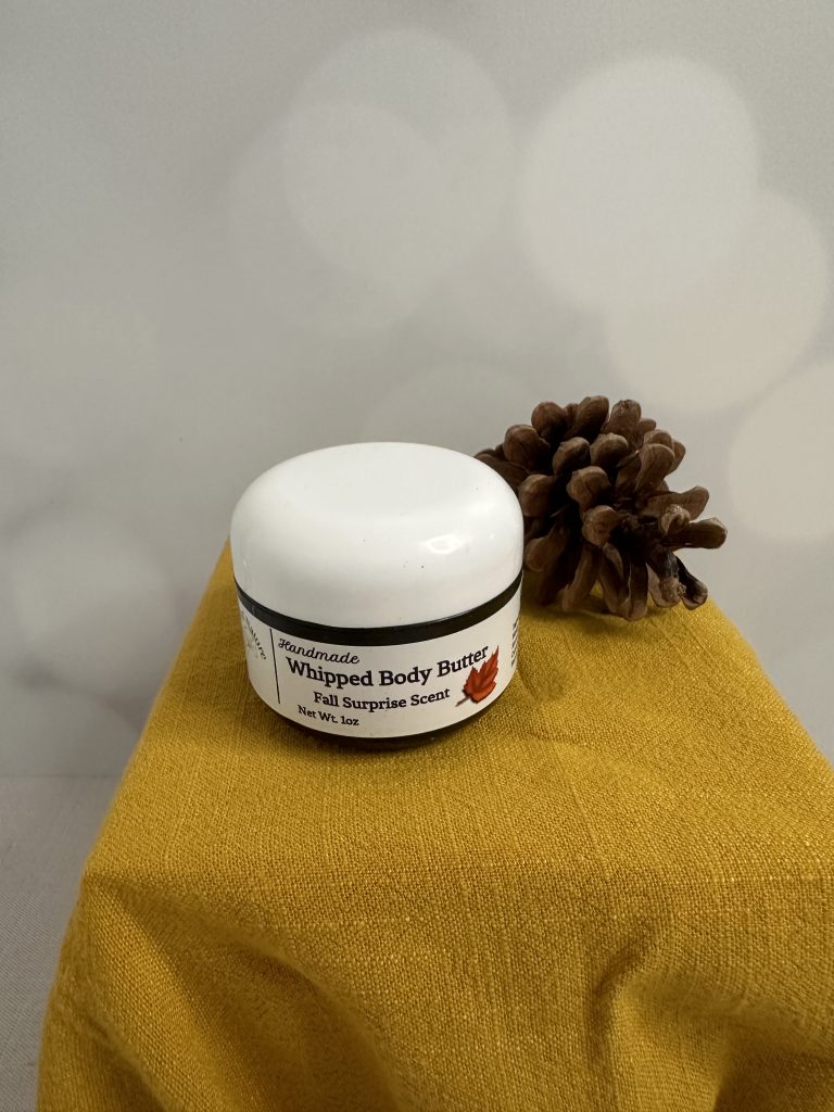 A whipped body butter in a black and white jar presented next to a pinecone on a box covered with a dark yellow blanket.
